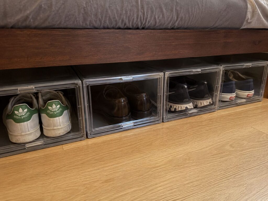 Row of clear drop-front shoe storage boxes under a bed showing sneakers and dress shoes organized by pair