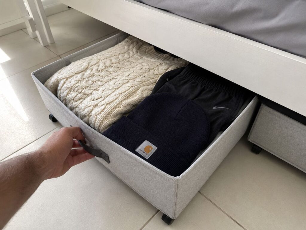 Low-profile rolling fabric storage drawer being pulled out from under a white bed frame in a small bedroom
