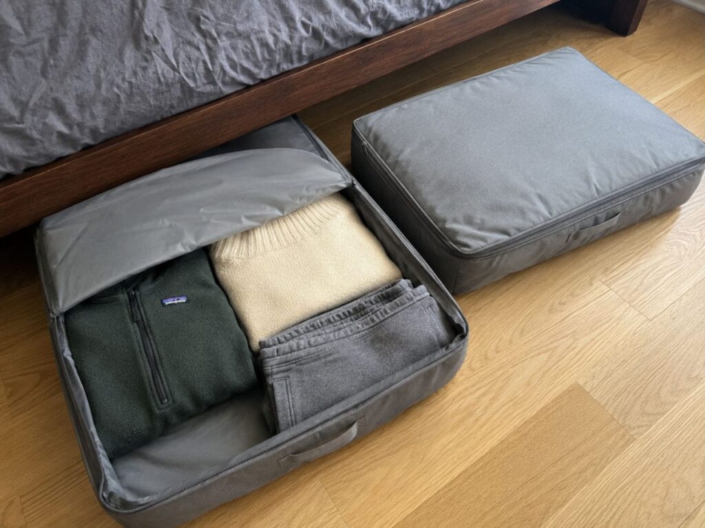 Two flat grey zip-up fabric storage bags filled with folded clothes next to a bed ready to slide under