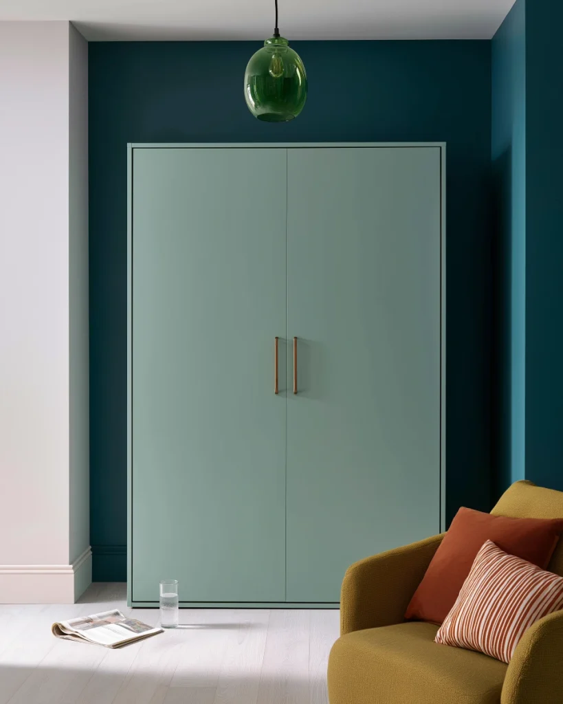 A closed wall bed cabinet in sage green joinery against a deep teal wall in a studio apartment, with a mustard linen chair beside it.